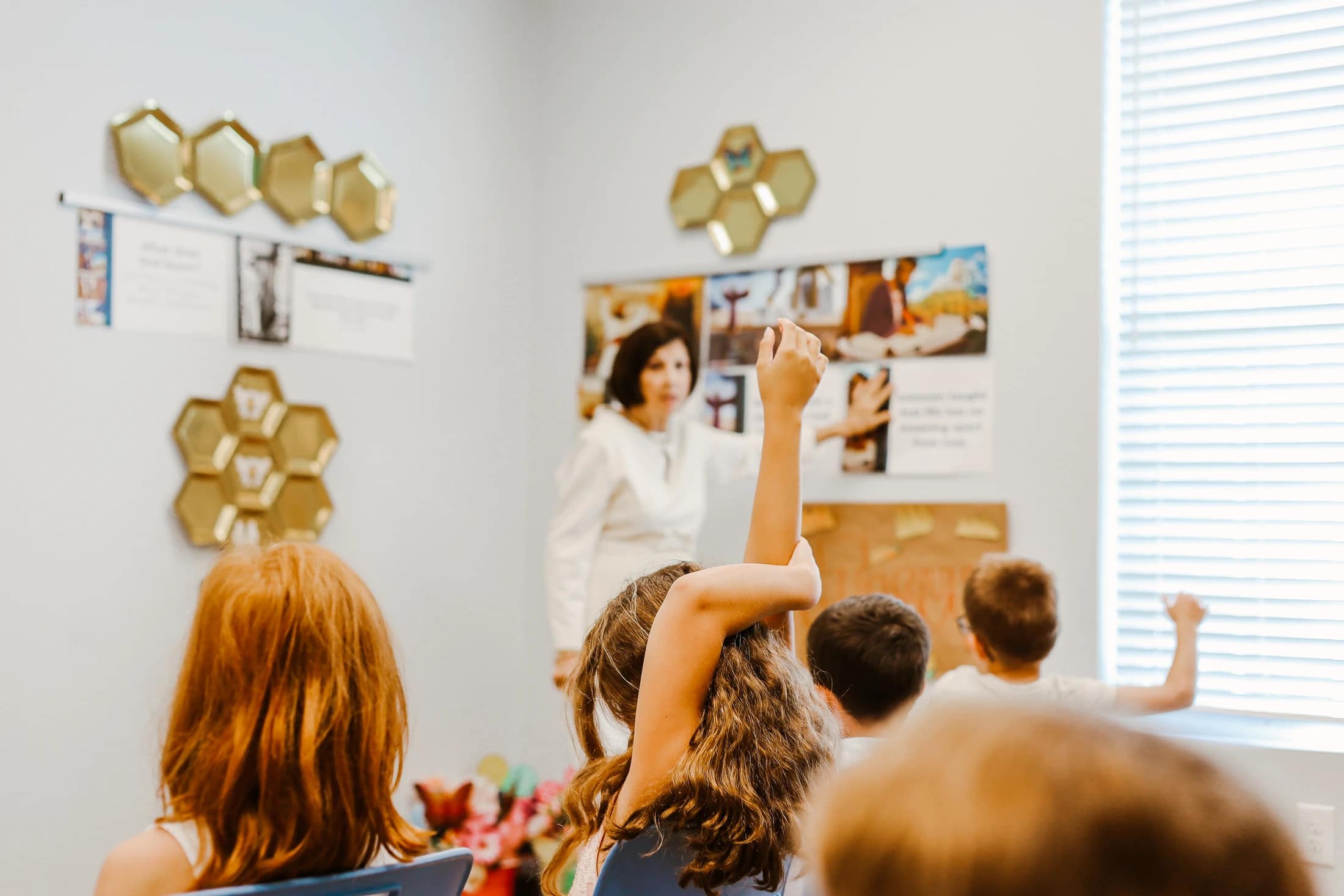 A view from behind a group of children in a classroom. One girl has her arm raised high to answer a question from the teacher at the front of the room.